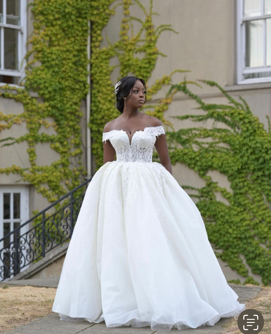 Woman in an elegant white wedding dress standing outdoors near a building with green ivy covering the walls.